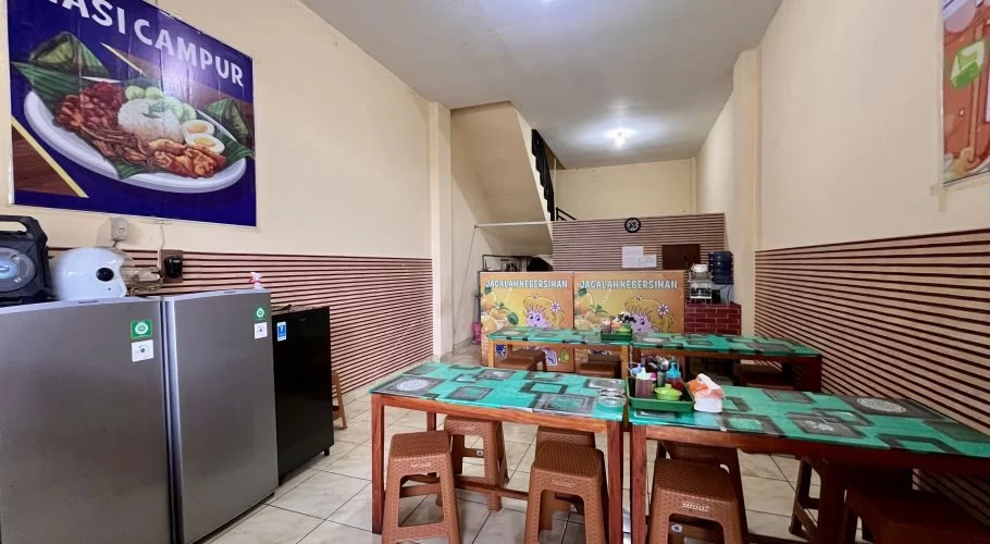 A clean, simple Indonesian eatery interior with cream walls, brown striped wainscoting, wooden tables, brown stools, and refrigerators.