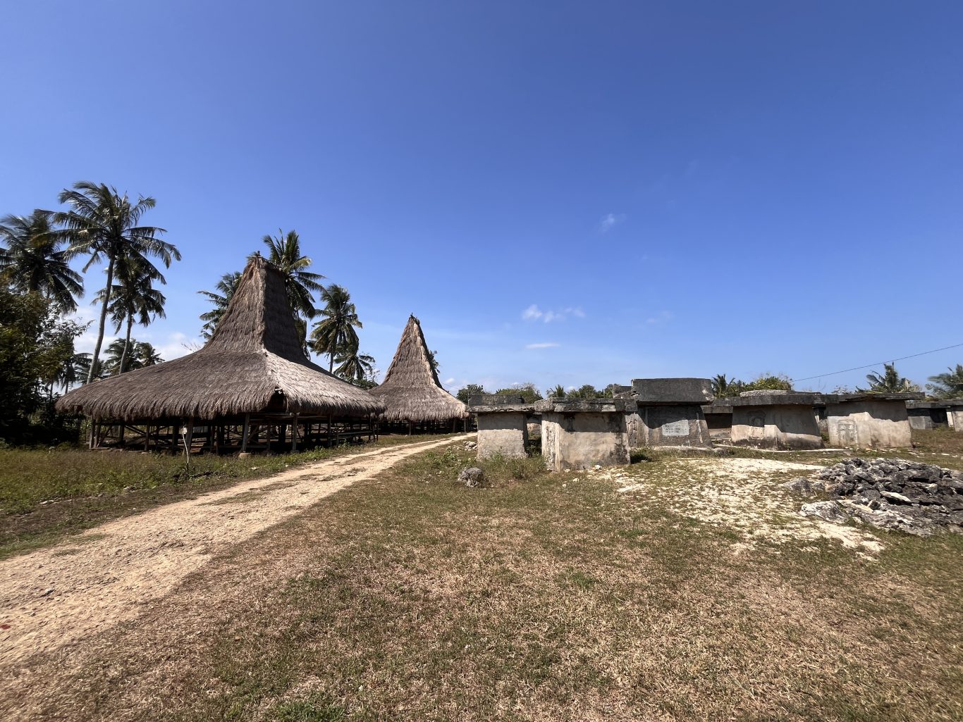 Two traditional Sumba houses with tall thatched roofs stand beside a dirt path, leading past large stone graves under a clear blue sky.