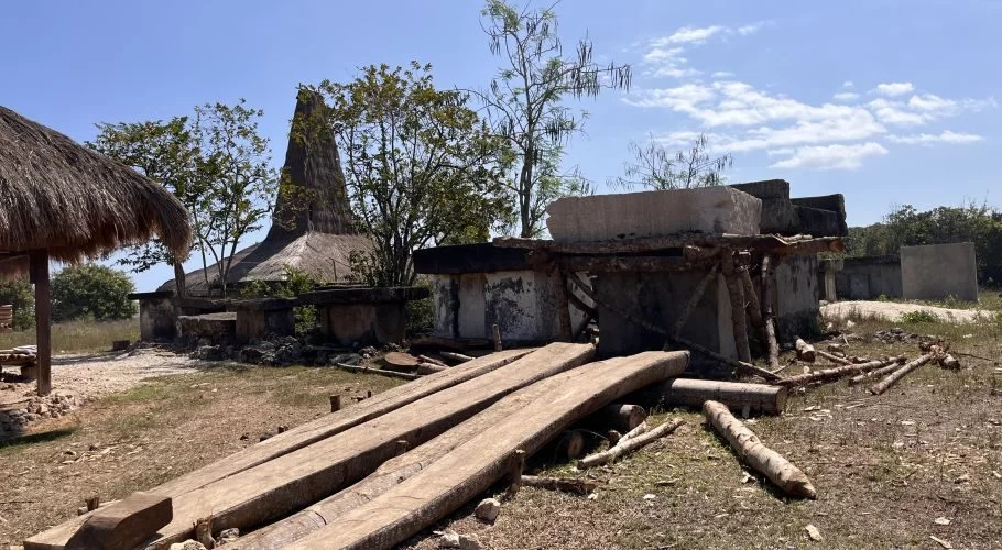 A traditional Sumba village features a tall thatched-roof house, large stone graves, and wooden planks on dry ground under a blue sky.