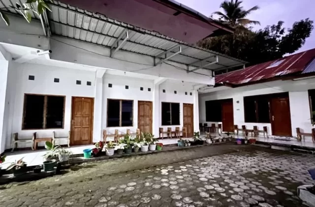 The exterior of a homestay features white rooms with brown wooden doors and dark windows, lining a paved courtyard with potted plants under a dark sky.