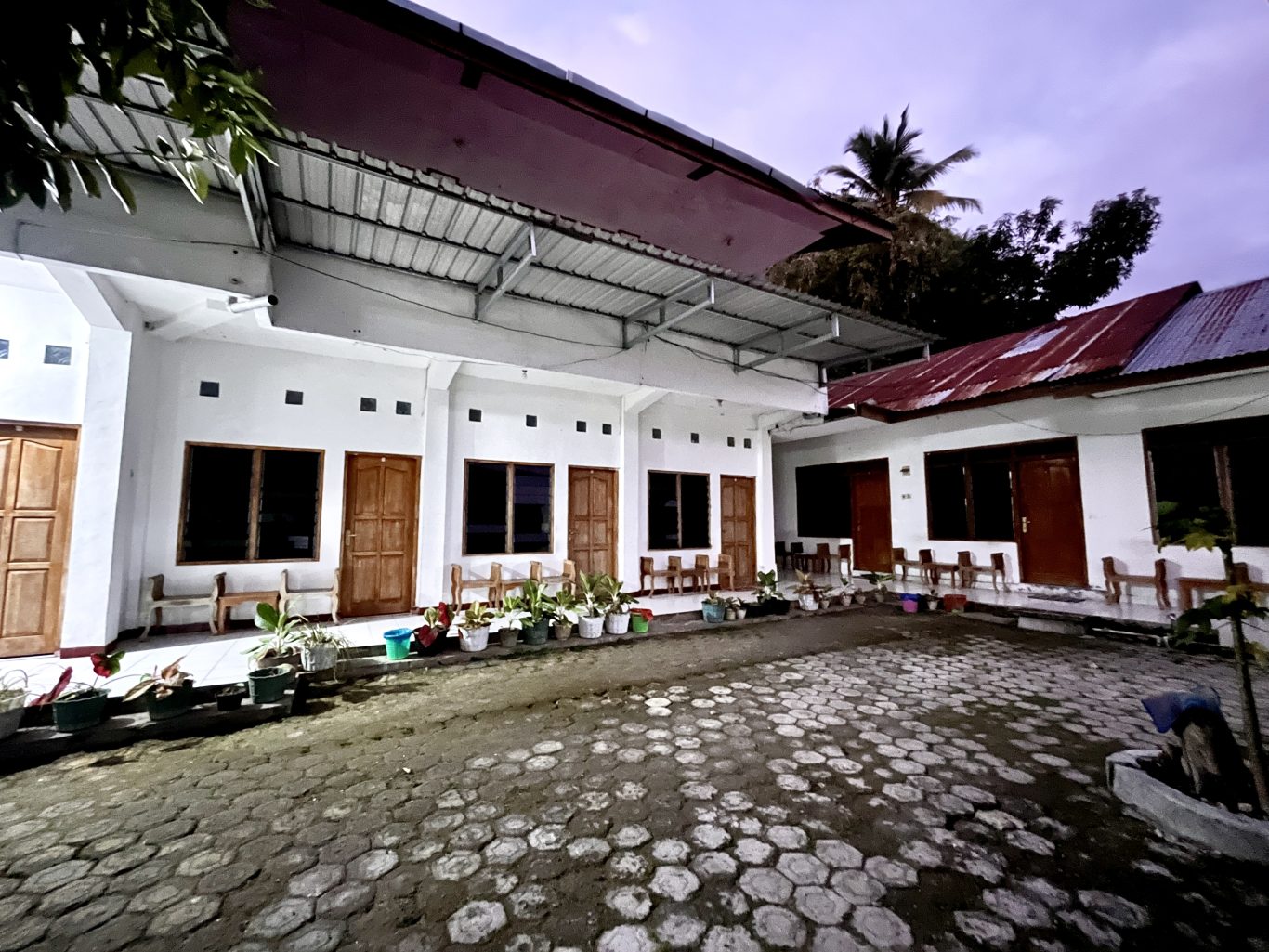 The exterior of a homestay features white rooms with brown wooden doors and dark windows, lining a paved courtyard with potted plants under a dark sky.