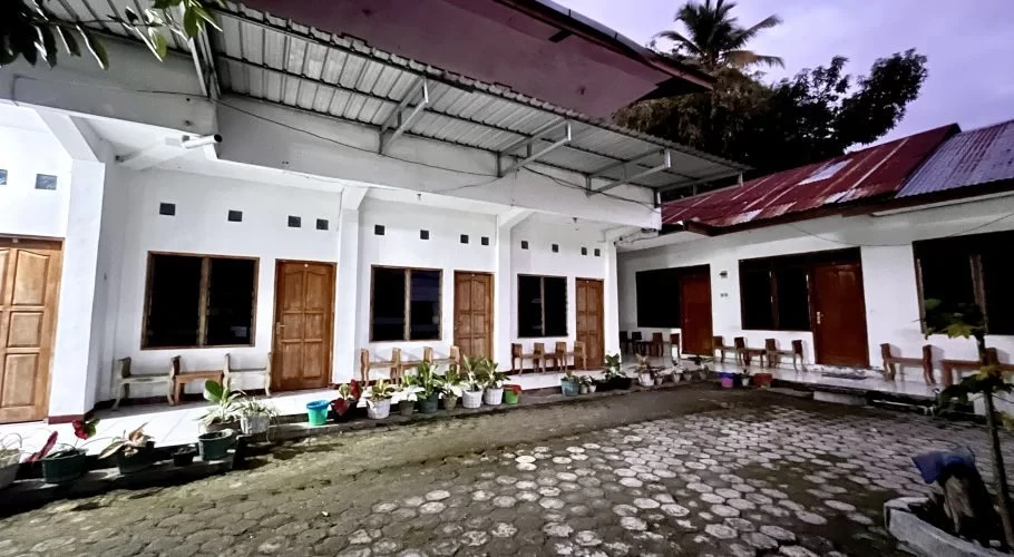 The exterior of a homestay features white rooms with brown wooden doors and dark windows, lining a paved courtyard with potted plants under a dark sky.
