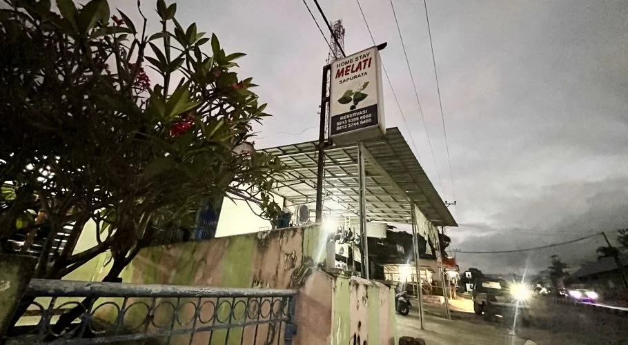 The Melati Homestay sign is visible above a building with a metal roof, framed by a leafy tree with red flowers, on a dark street with car headlights.