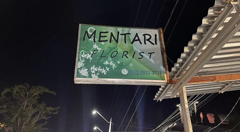 A green and white sign for "MENTARI FLORIST" is illuminated at night, showing a phone number and floral patterns, attached to a building with a corrugated metal roof.
