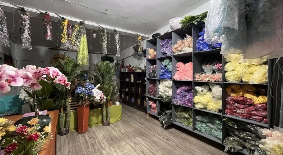 A florist shop interior displays shelves packed with colorful artificial flowers in plastic bags, alongside large pink orchids and green decorative plants.