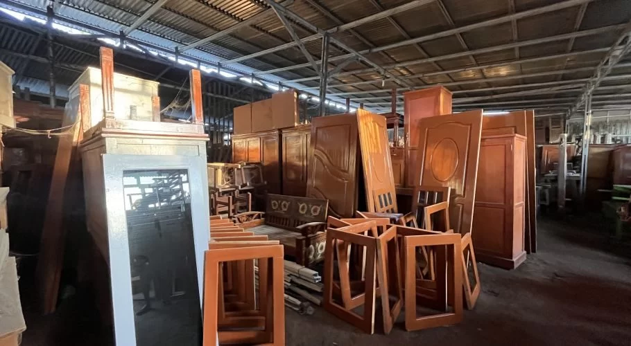 Visible is a large carpentry workshop interior in Waingapu, East Sumba, filled with numerous reddish-brown wooden doors, cupboards, and furniture frames under a corrugated metal roof.