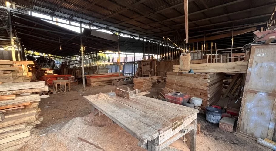 A rustic carpentry workshop interior features large stacks of raw timber, finished wood pieces, and a saw table covered in sawdust under a metal roof.