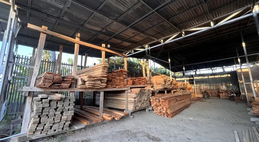 Numerous stacks of brown and reddish-brown lumber, including planks and beams, are organized within a large, open-sided carpentry workshop shed.