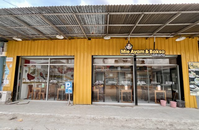 The exterior of Mie Ayam & Bakso Jakarta, a yellow restaurant building with large glass windows, a corrugated metal roof, and a blue parking sign on the sidewalk in Waikabubak.