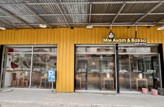 The exterior of Mie Ayam & Bakso Jakarta, a yellow restaurant building with large glass windows, a corrugated metal roof, and a blue parking sign on the sidewalk in Waikabubak.