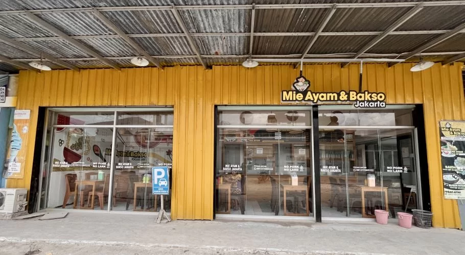 The exterior of Mie Ayam & Bakso Jakarta, a yellow restaurant building with large glass windows, a corrugated metal roof, and a blue parking sign on the sidewalk in Waikabubak.