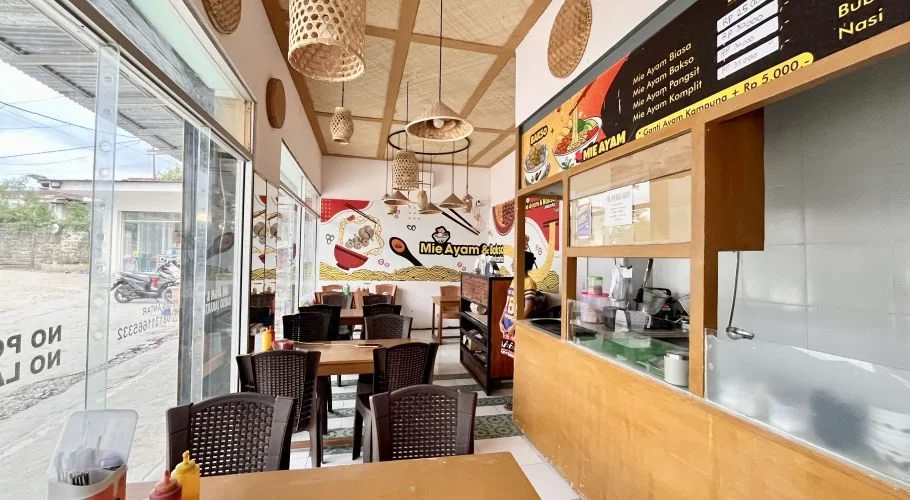 Restaurant interior with wooden tables, dark chairs, and a colorful mural depicting food on a white wall. Woven bamboo light fixtures hang from the ceiling, and large windows show a street outside.