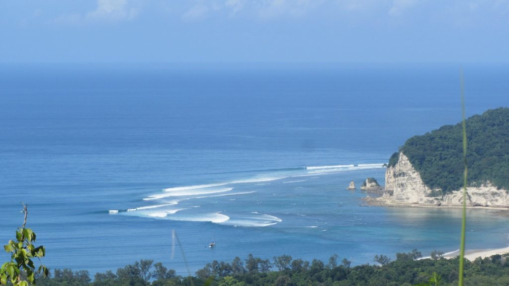 Blue ocean waves break towards a white cliff and a green forested hill on a sunny day, with a small boat anchored in the bay.