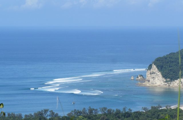 Blue ocean waves break towards a white cliff and a green forested hill on a sunny day, with a small boat anchored in the bay.