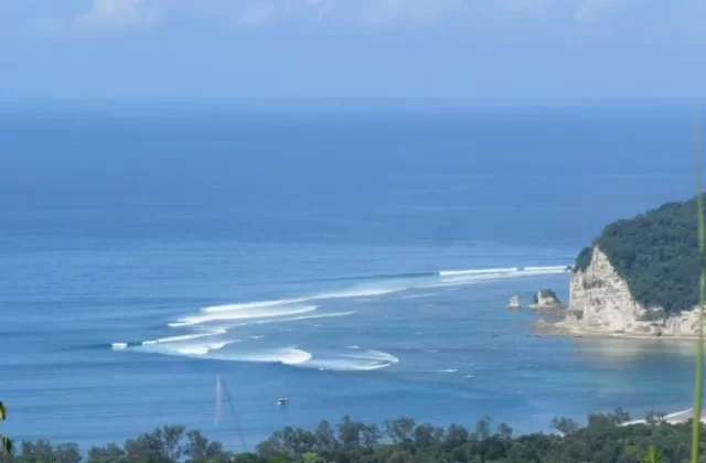 Blue ocean waves break towards a white cliff and a green forested hill on a sunny day, with a small boat anchored in the bay.
