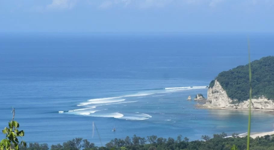 Blue ocean waves break towards a white cliff and a green forested hill on a sunny day, with a small boat anchored in the bay.