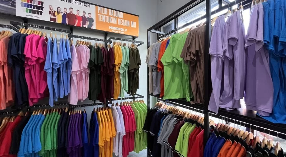 Inside Monopoly Printing, numerous vibrant t-shirts in various solid colors like pink, blue, green, and yellow hang neatly on black display racks.