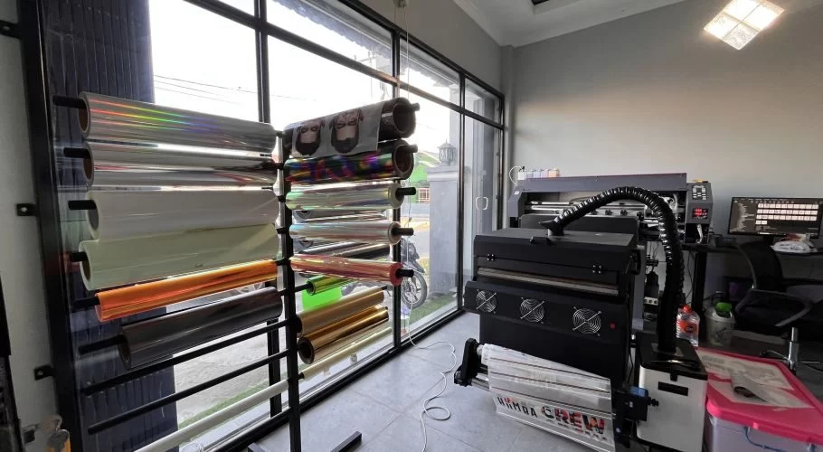 Inside Monopoly Printing, a rack holds colorful vinyl rolls for custom designs, positioned near a large black printing machine and a computer workstation.