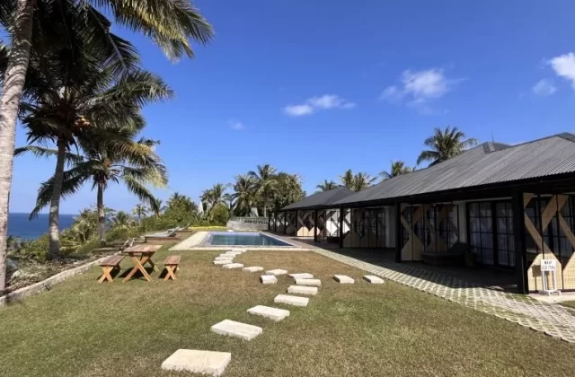 A sunny coastal scene features two white buildings with dark roofs, a swimming pool, green lawn, palm trees, and the blue ocean under a clear sky.