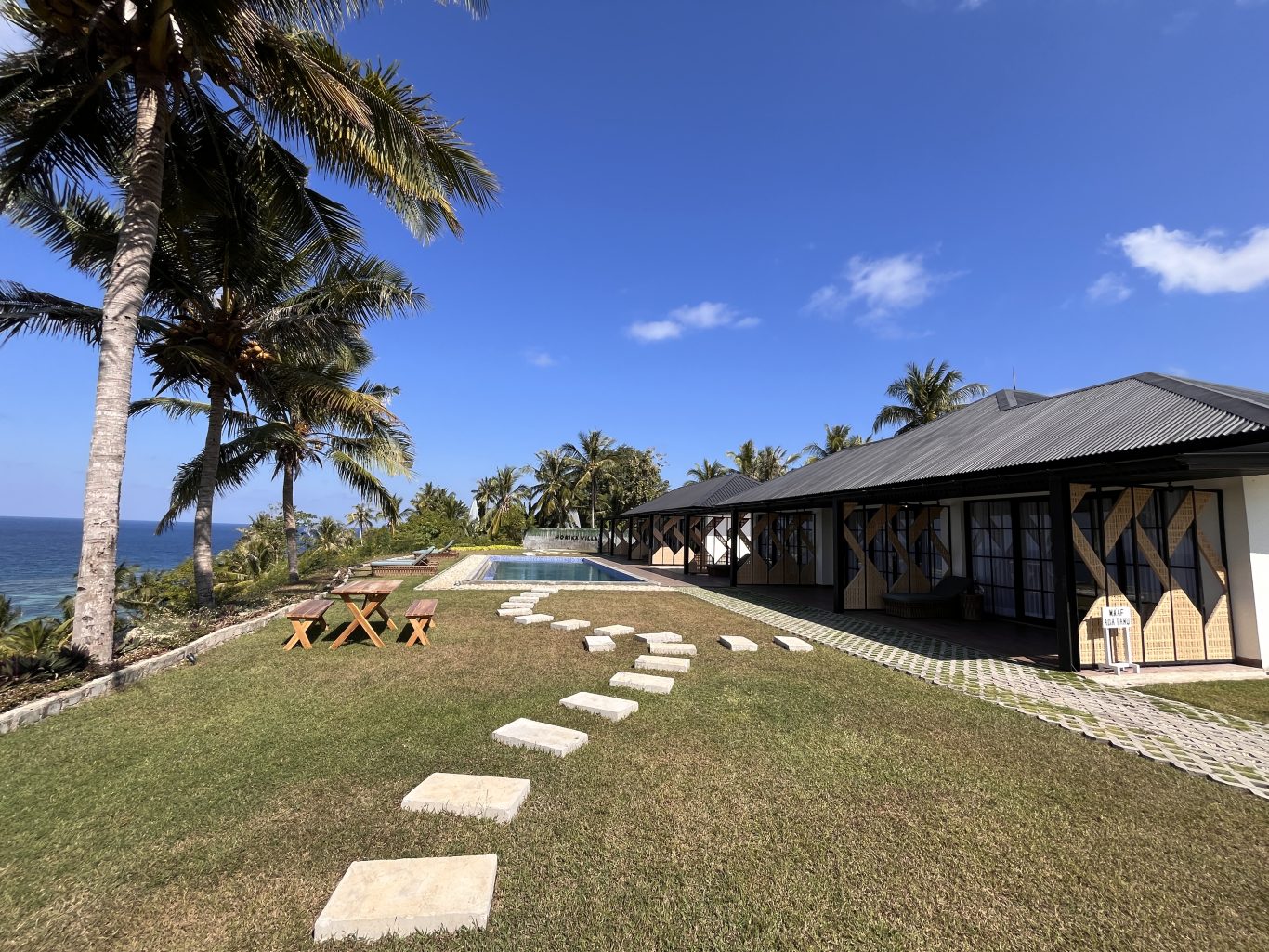 A sunny coastal scene features two white buildings with dark roofs, a swimming pool, green lawn, palm trees, and the blue ocean under a clear sky.