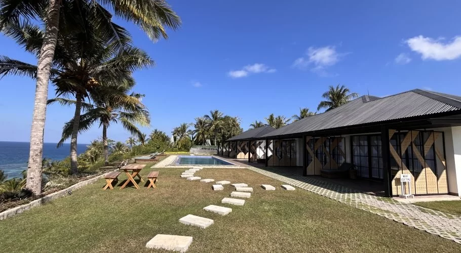 A sunny coastal scene features two white buildings with dark roofs, a swimming pool, green lawn, palm trees, and the blue ocean under a clear sky.