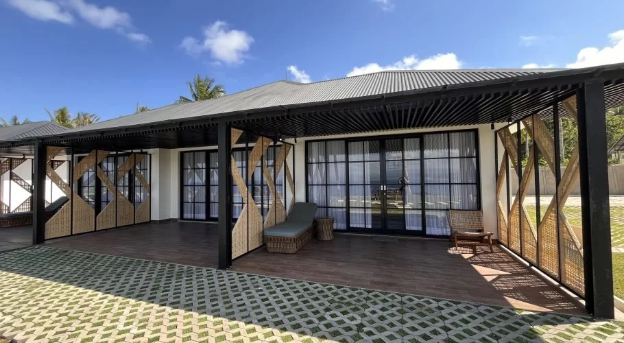 Modern white cottage with a dark roof, large glass doors, and a covered patio featuring bamboo screens and a chaise lounge. A grass grid is in the foreground under a blue sky.