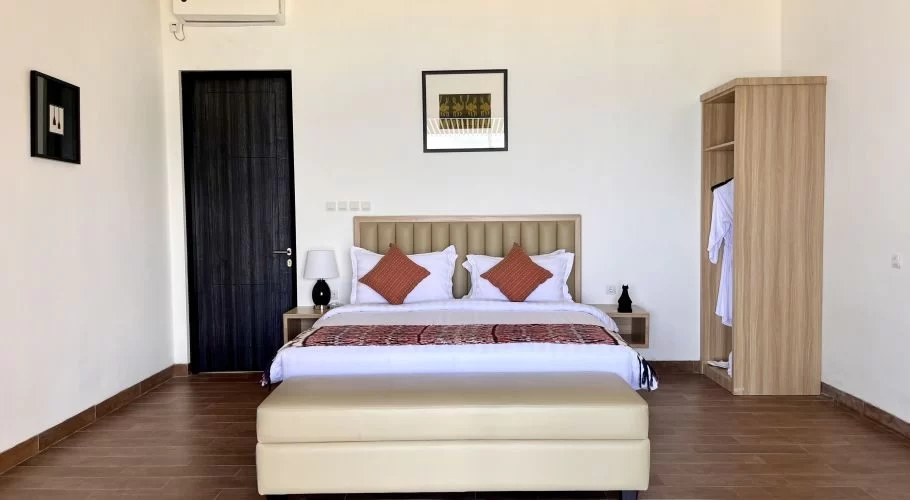 A modern bedroom at Morika Cottage features a large bed with a beige headboard, white linens, and a patterned throw, flanked by a dark door and an open wardrobe.
