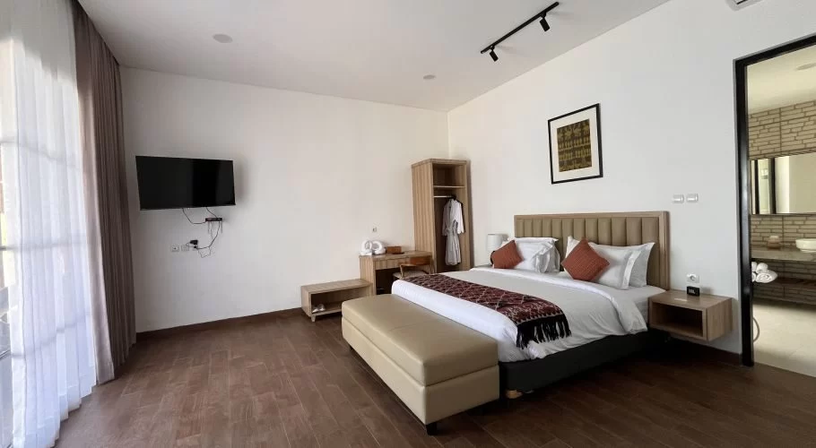 A modern bedroom features a large bed with white linens and orange pillows, a beige bench, a wall-mounted TV, and a wooden wardrobe. A doorway on the right leads to a stylish bathroom.