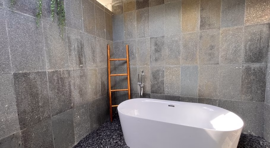 A modern white freestanding bathtub sits on black pebbles in an outdoor bathroom with dark grey stone tiled walls, a wooden ladder, and green vines.