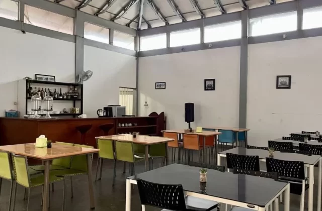 A brightly lit hotel dining area features a wooden bar with drinks, various tables with colorful chairs, white walls, and a high ceiling with windows.