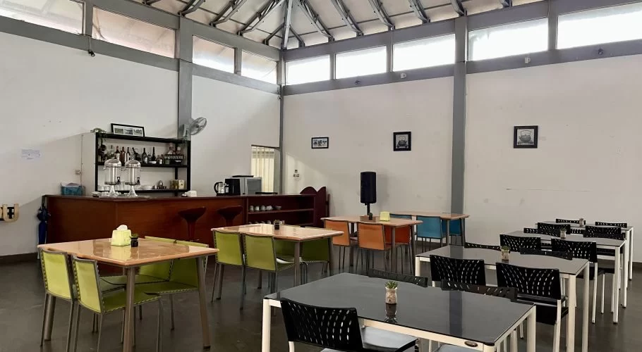 A brightly lit hotel dining area features a wooden bar with drinks, various tables with colorful chairs, white walls, and a high ceiling with windows.