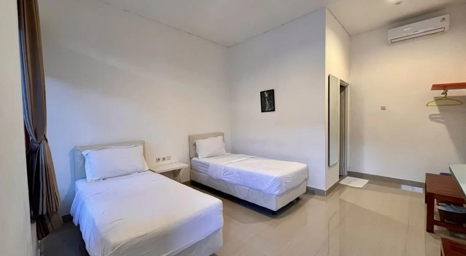 A twin hotel room features two single beds with white bedding, white walls, a tiled floor, brown curtains, and an air conditioner.