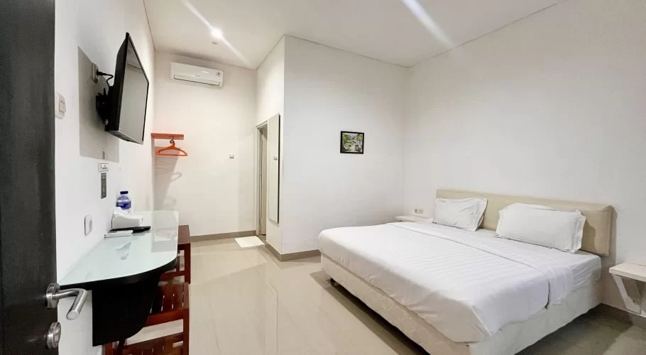 A clean, modern hotel room features a large bed with white linens, a wall-mounted TV, a desk, and an air conditioner.