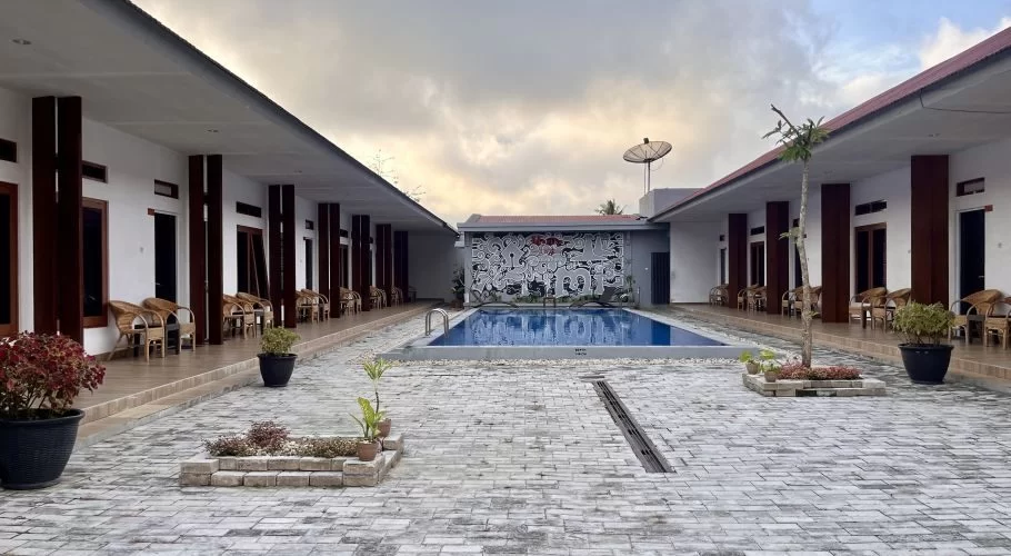 A hotel courtyard features a blue swimming pool flanked by two white buildings with dark brown accents and rattan chairs, under an overcast sky.