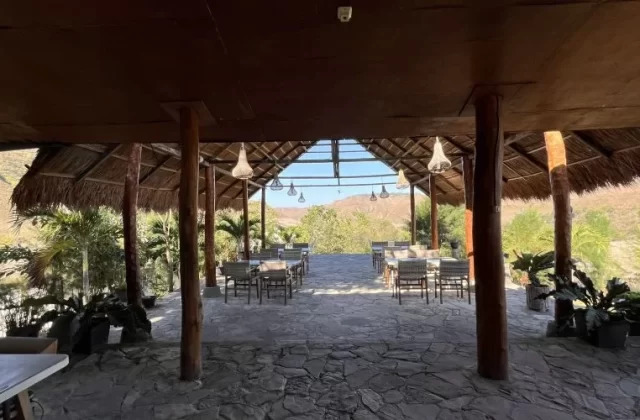 An open-air restaurant with a thatched roof and wooden pillars features dining tables on a stone floor, overlooking dry hills and green foliage.