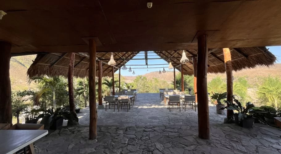 An open-air restaurant with a thatched roof and wooden pillars features dining tables on a stone floor, overlooking dry hills and green foliage.