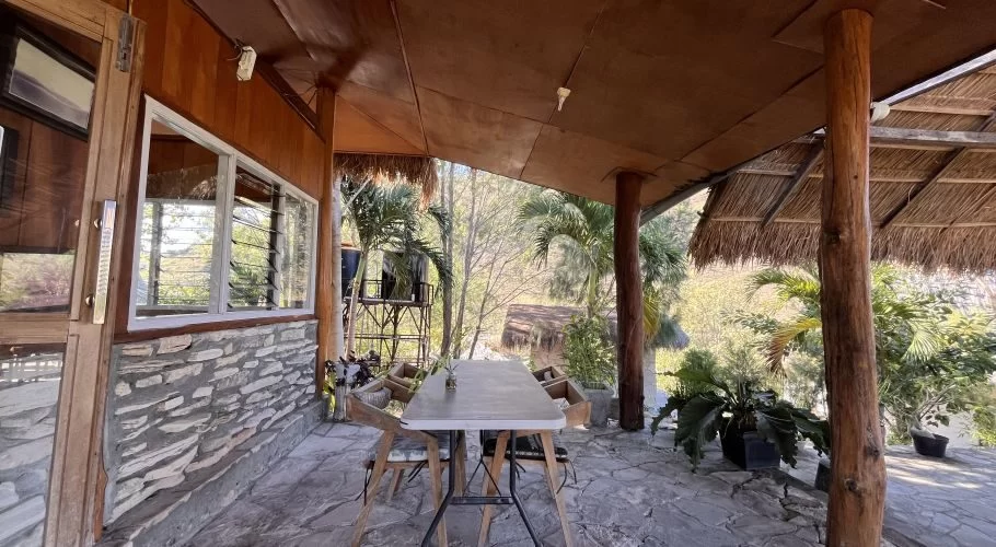 Outdoor dining patio at Morinda Restaurant with a table and chairs on a stone floor, under a wooden and thatched roof, overlooking lush trees.