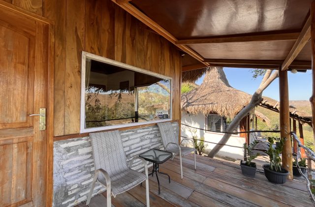 A wooden villa balcony with a door, large window, two grey chairs, and a table, overlooks a thatched-roof building and distant hills.