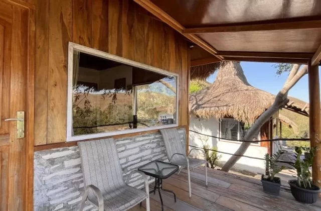 A wooden villa balcony with a door, large window, two grey chairs, and a table, overlooks a thatched-roof building and distant hills.
