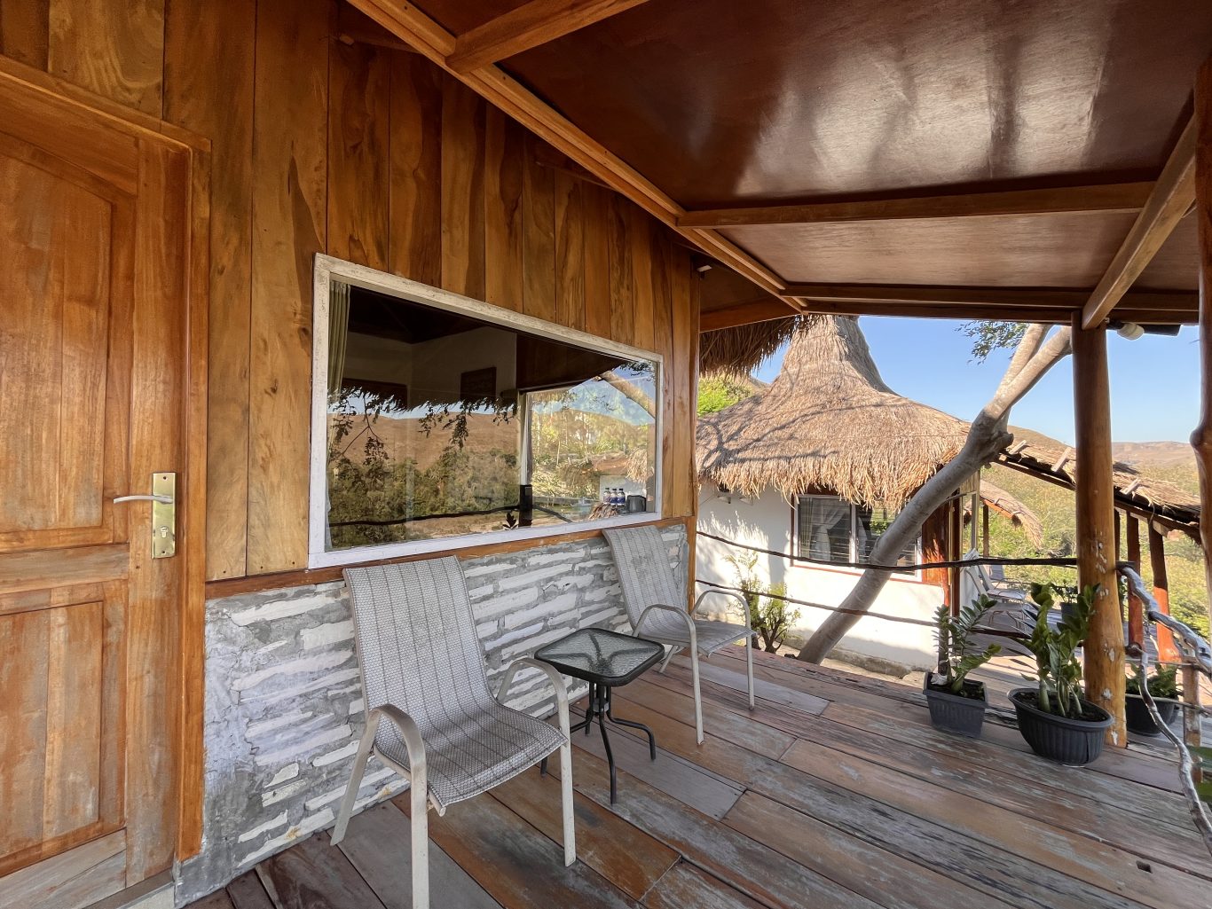 A wooden villa balcony with a door, large window, two grey chairs, and a table, overlooks a thatched-roof building and distant hills.