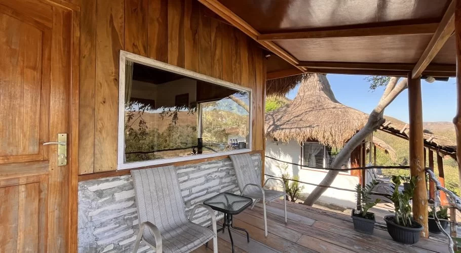 A wooden villa balcony with a door, large window, two grey chairs, and a table, overlooks a thatched-roof building and distant hills.