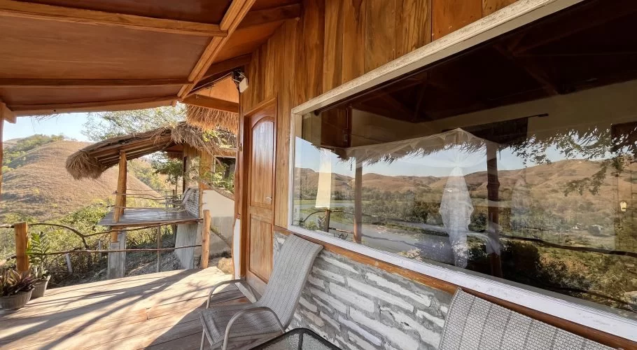 A wooden villa balcony with a large window overlooks golden rolling hills and the Lambanapu River under a clear blue sky.
