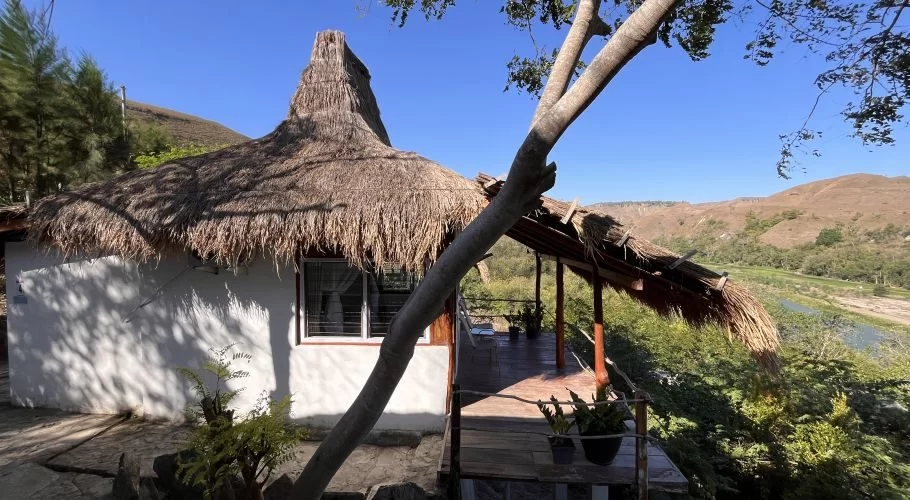 A white villa with a tall thatched roof and a wooden deck overlooks a winding river and dry, rolling hills under a clear blue sky.