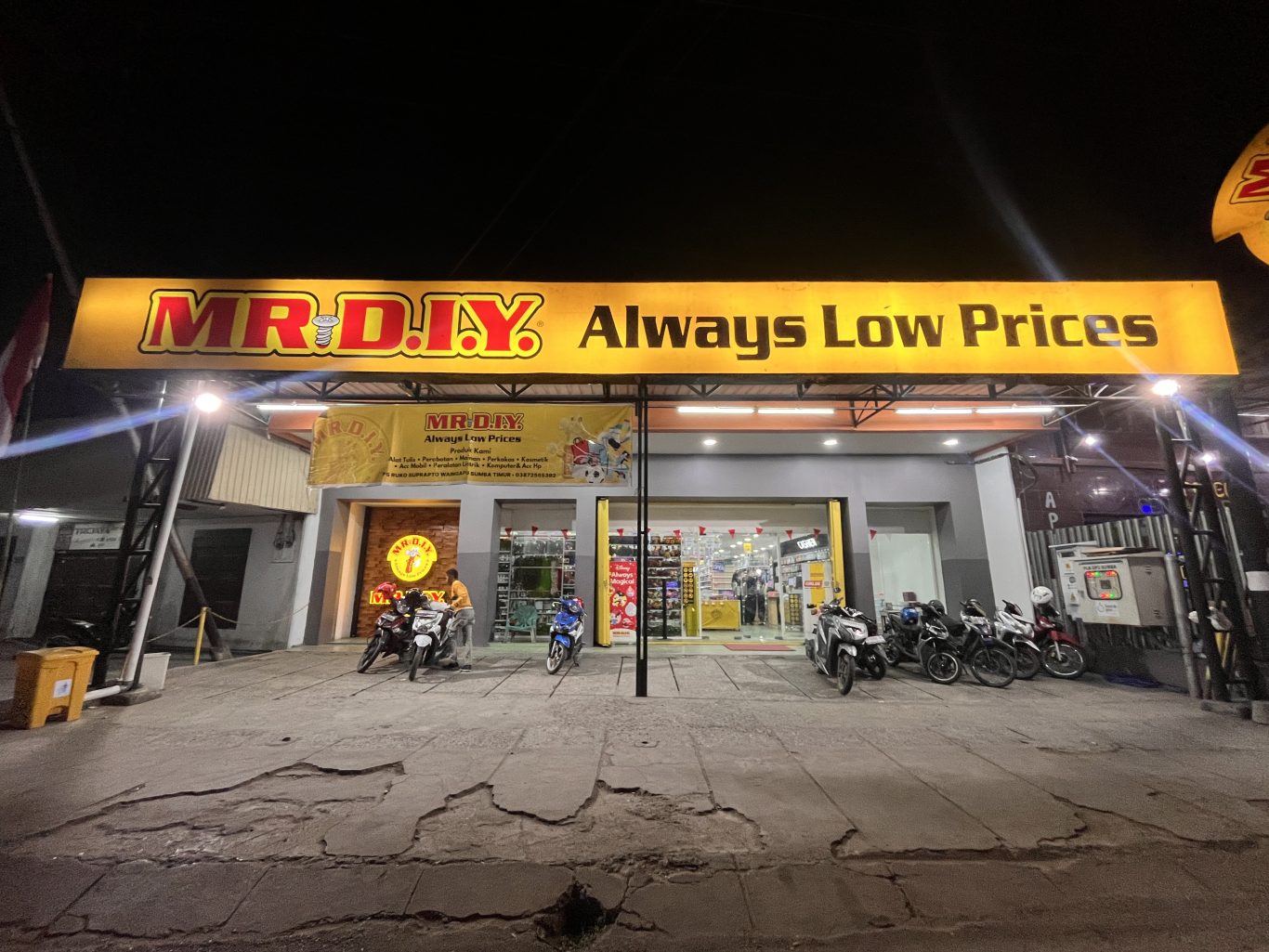 The brightly lit exterior of a MR. DIY store at night, featuring a large yellow sign and several motorcycles parked in front.