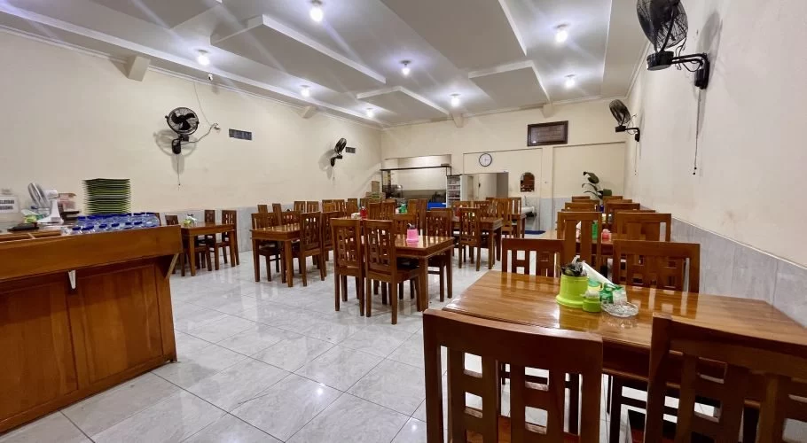 A clean, spacious restaurant interior with many wooden tables and chairs, light walls, a tiled floor, and wall-mounted fans.