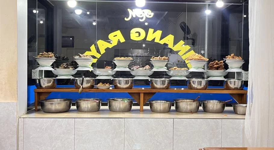 A brightly lit restaurant window showcases a tiered display of various Indonesian dishes in white and silver bowls, with yellow lettering on the dark glass.