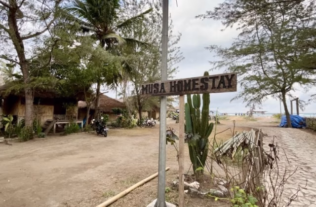 A wooden "MUSA HOMESTAY" sign stands near traditional thatched-roof bungalows and palm trees, with a sandy path leading to a beach and ocean under a cloudy sky.