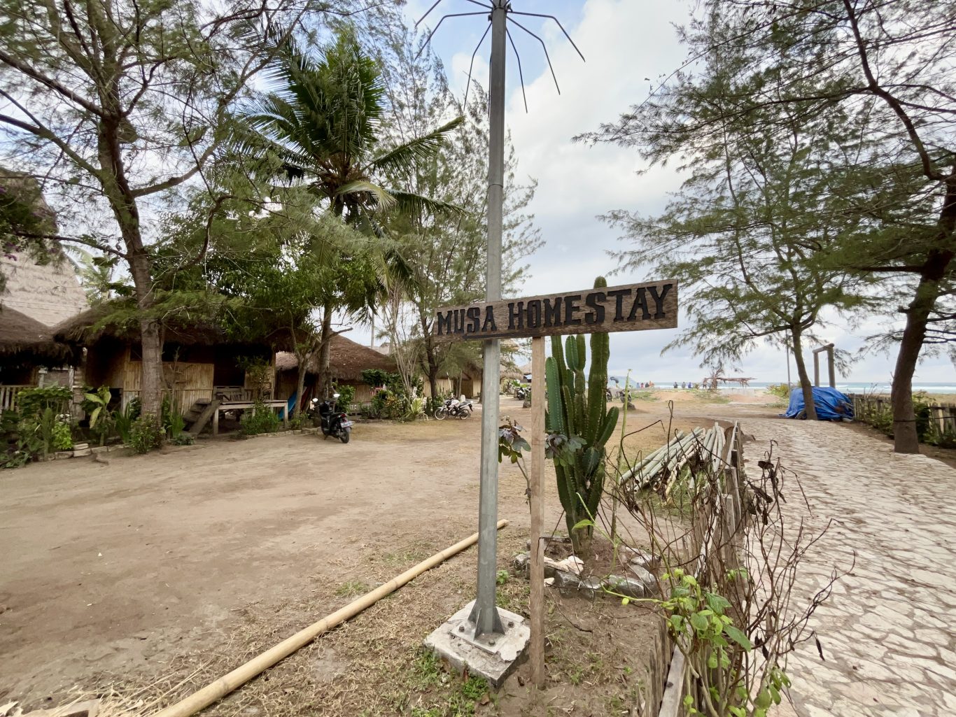 A wooden "MUSA HOMESTAY" sign stands near traditional thatched-roof bungalows and palm trees, with a sandy path leading to a beach and ocean under a cloudy sky.
