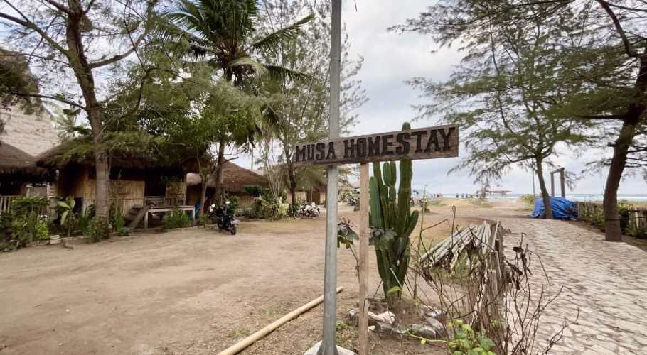 A wooden "MUSA HOMESTAY" sign stands near traditional thatched-roof bungalows and palm trees, with a sandy path leading to a beach and ocean under a cloudy sky.