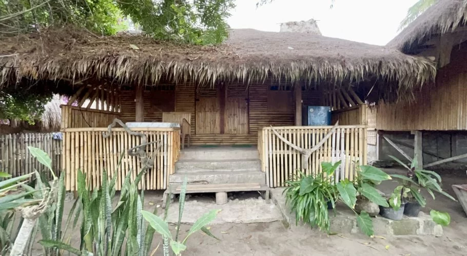 A traditional Sumba-style homestay building features a thick thatched roof, bamboo walls, concrete steps, and a small bamboo-railed balcony, surrounded by green plants.